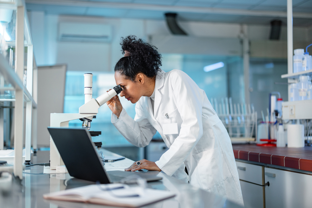 Scientist wearing a lab coat, looking under microscope while using laptop in a laboratory